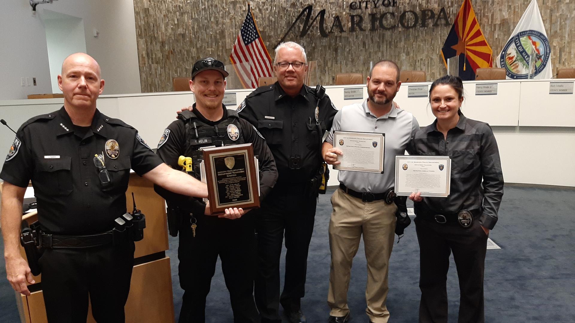 Five police officers stand proudly with awards in a council chamber, in front of flags and a 