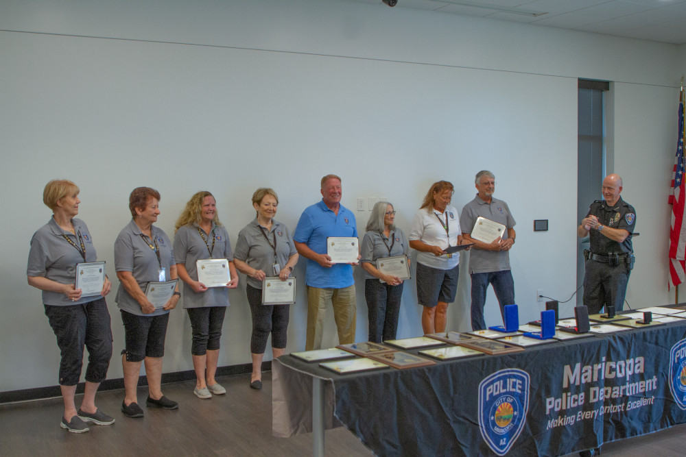 Eight people stand holding certificates in a room, with the Maricopa Police Department logo visible on a table. A police officer applauds them.
