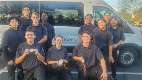 A group of smiling young adults in matching uniforms stand in front of a white van. Three in front hold medals. The scene is bright and conveys achievement.
