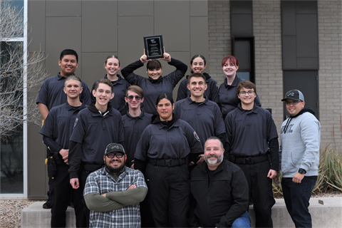 A group of 13 people, mostly in matching dark uniforms, pose happily outside a building. One holds a plaque overhead, celebrating a shared achievement.
