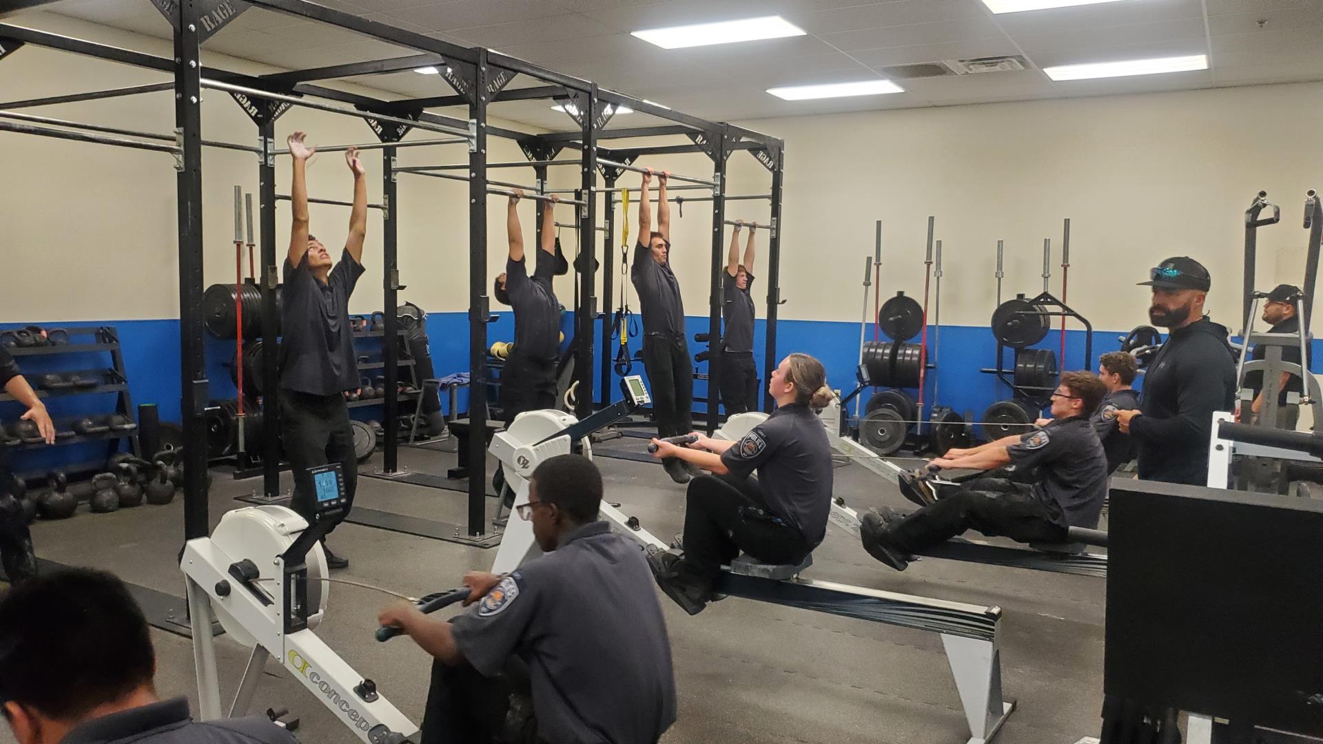 A group of uniformed individuals exercise in a gym. Some are doing pull-ups, while others use rowing machines. The atmosphere is focused and energetic.