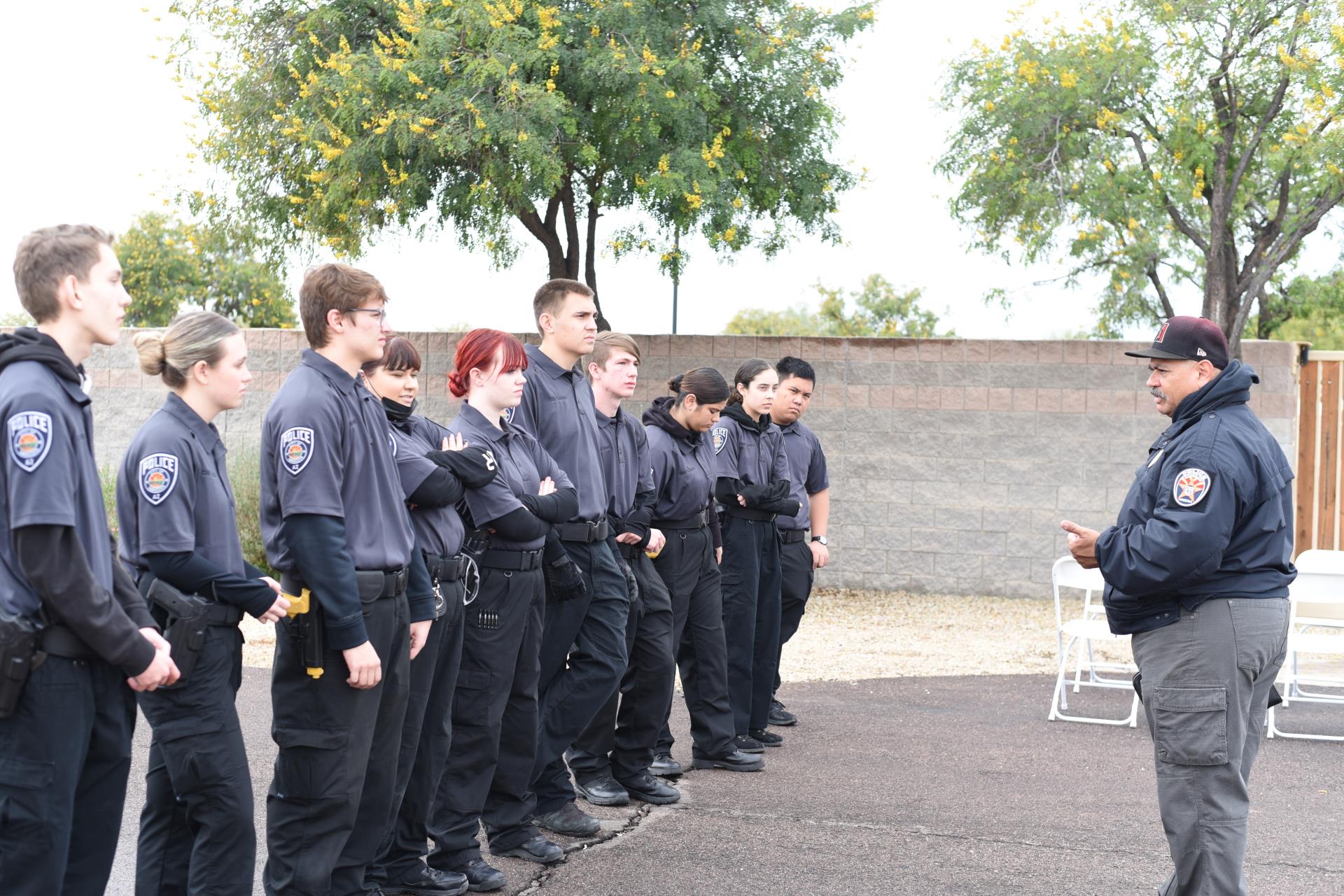 A group of uniformed cadets stands in line attentively facing an instructor outdoors. The setting is calm, with trees and a brick wall in the background.