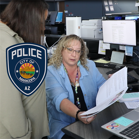 A woman showing documents to another person at an office desk, surrounded by computer monitors and paperwork. 