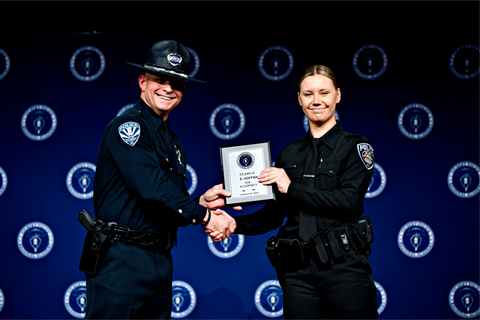 Two police officers in uniform shake hands on stage. One officer holds a plaque. Both smile against a backdrop with a police emblem pattern.