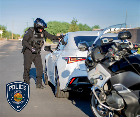 Police officer in black uniform and helmet approaches white car on a sunny street. Police motorcycle in foreground; City of Maricopa emblem visible.