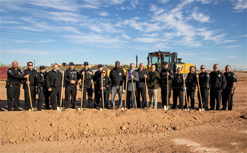 A group of people, including police officers and civilians, stand on a dirt construction site holding shovels, with a bulldozer behind them on a clear day.