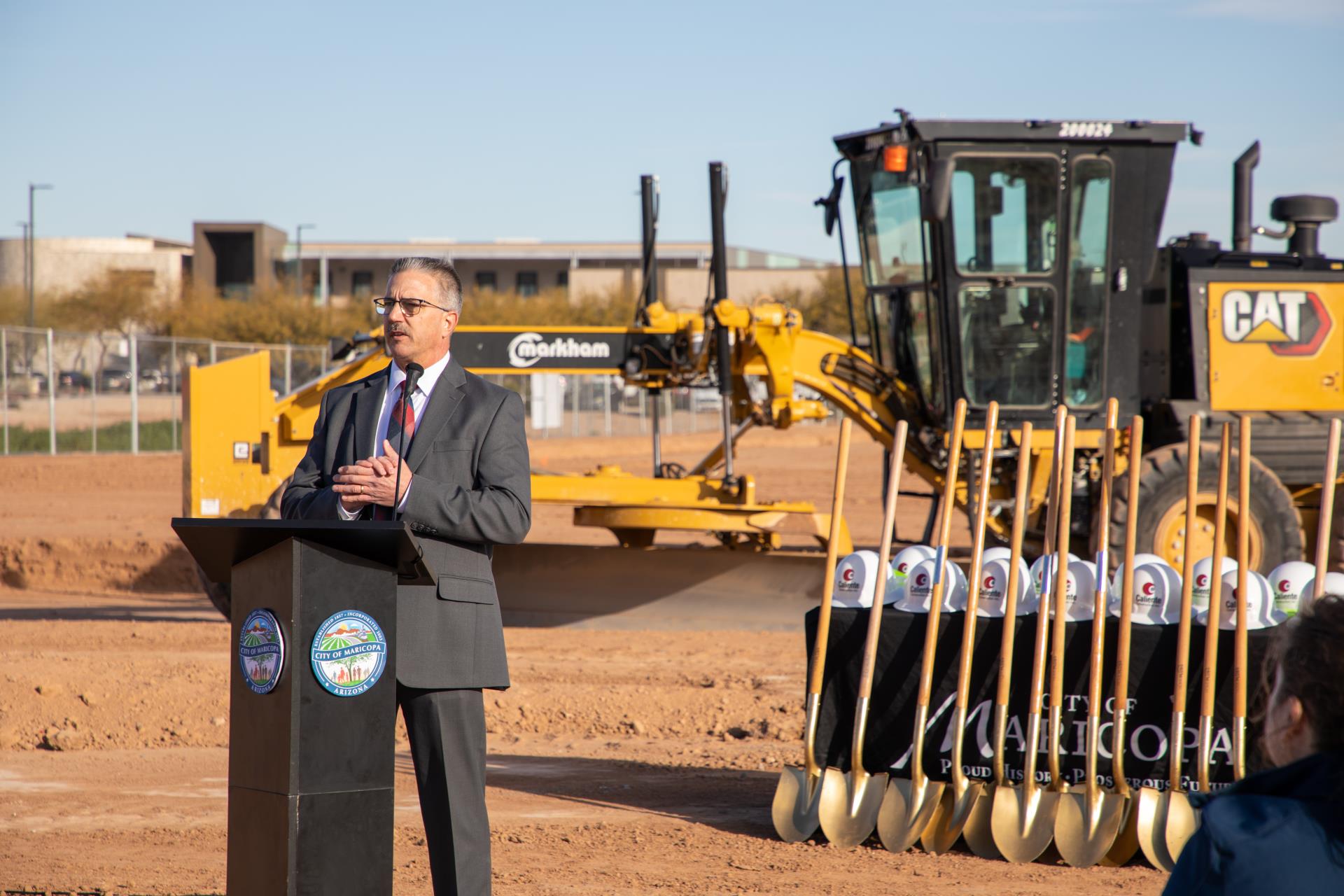 A man in a suit speaks at a podium at a groundbreaking ceremony. Behind him are a row of gold shovels and a large construction vehicle, set against a clear blue sky.