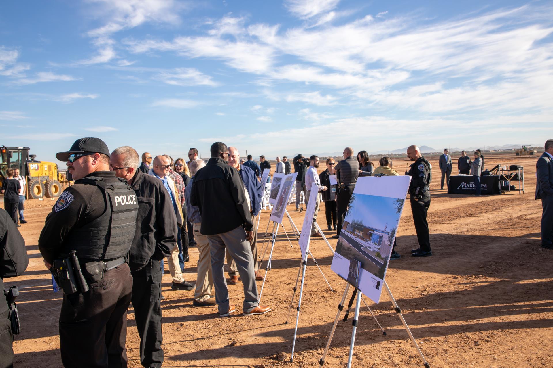 Outdoor event on a sunny day with blue skies. People, including police officers, are gathered around display boards on a dirt surface, creating a focused, professional atmosphere.