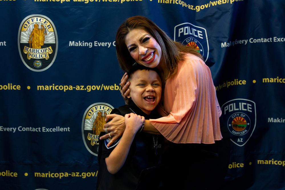 A joyful woman in a peach blouse hugs a smiling boy in a police uniform. They stand before a backdrop with Maricopa Police logos and the website URL.