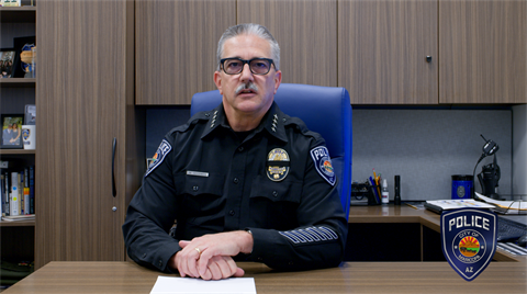A police officer sits at a desk in an office, wearing a black uniform with badges on the sleeves and chest. He has a serious expression, with books on shelves in the background. A police department emblem is visible in the bottom right corner.