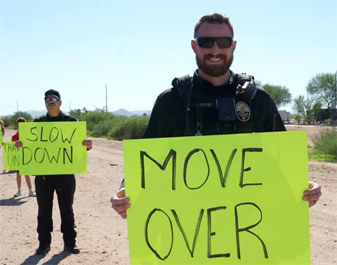 Police officer smiling, holding a bright yellow sign reading 