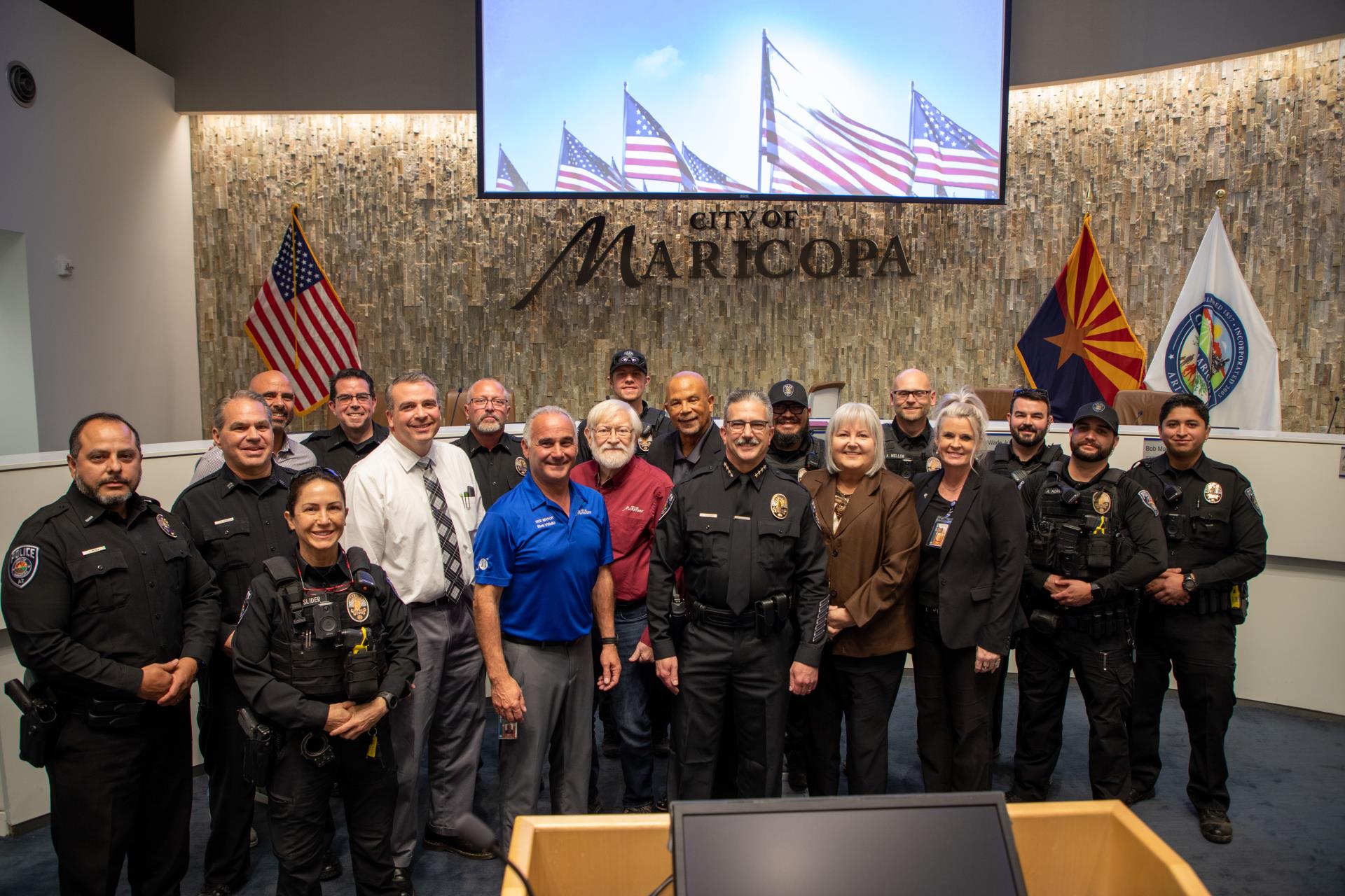 A group of police officers and city officials pose together in front of flags and a 