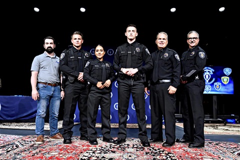 A group of five uniformed police officers and one civilian stand confidently on stage, framed by a backdrop of police insignias, conveying pride and unity.