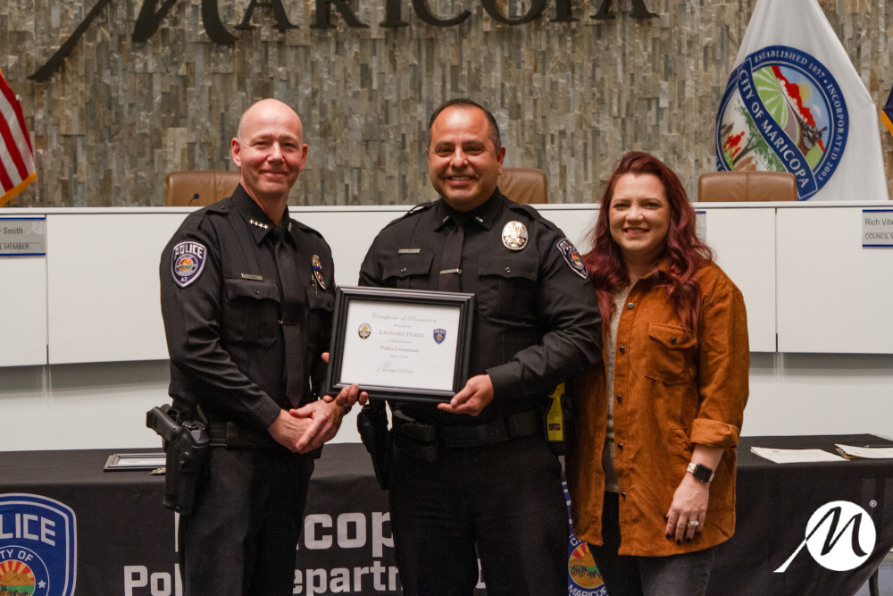 Three people stand inside a formal room. Two police officers in uniform hold a framed certificate, smiling proudly. A woman in casual attire stands beside them.