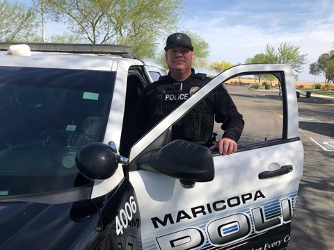 A police officer stands beside a Maricopa police vehicle with an open door. He is wearing a uniform and cap, and the scene is outdoors with trees and a blue sky.