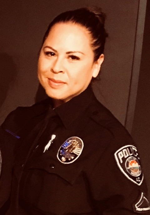 Female police officer in uniform, smiling with a badge and shoulder patch visible. The setting is neutral, conveying professionalism and confidence.