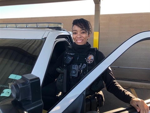 Smiling police officer stands beside an open patrol car door, wearing tactical gear, conveying confidence and approachability. Sunny outdoor setting.