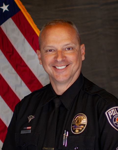Man in police uniform smiling, standing in front of the American flag. Wearing badges and insignia, conveys professionalism and dedication.