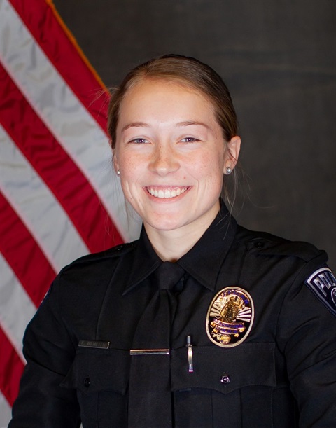 Smiling police officer in uniform stands in front of an American flag, conveying a sense of pride and service. The badge and insignia are visible.