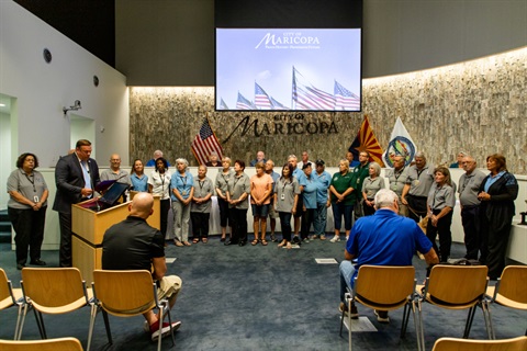 A group of people stands in a semicircle in a room with rows of chairs and a podium. Flags and 