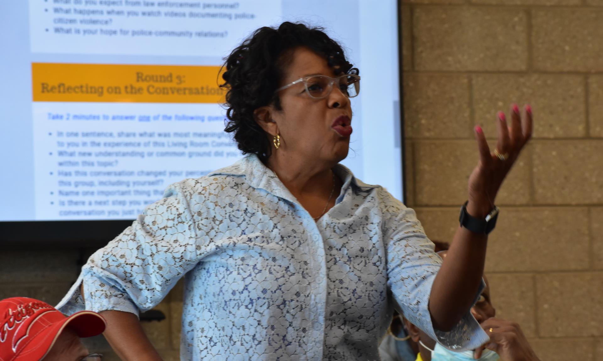 Woman passionately speaks in a discussion setting, gesturing with her hand. She's in front of a presentation, conveying engagement and leadership.