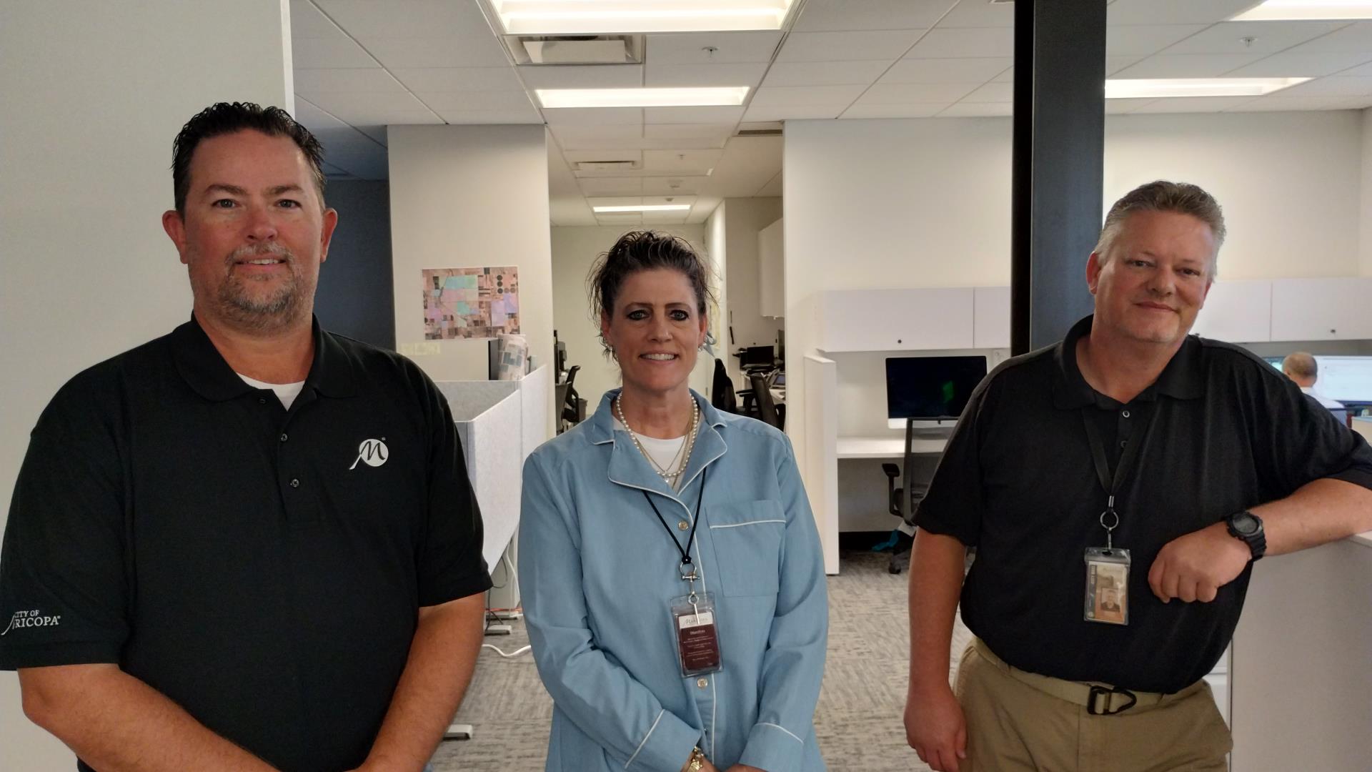 Three people stand in an office corridor, smiling warmly. They are dressed in business casual attire, conveying a friendly and professional atmosphere.