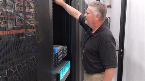 A man in a black shirt manages server equipment in a data center, standing next to a server rack filled with cables. The atmosphere is professional and focused.
