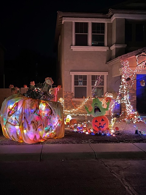 A house decorated for Halloween at night, featuring glowing pumpkins, spooky figures, colorful lights, and an oversized lit pumpkin in the yard.