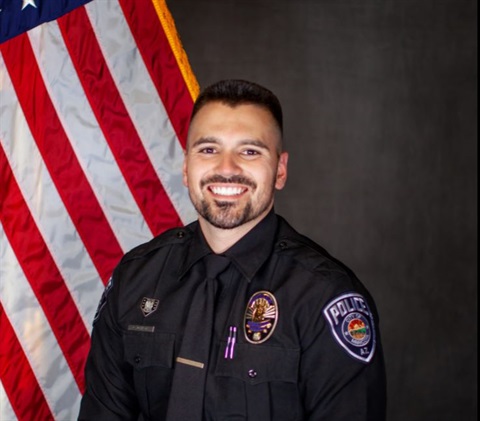 Smiling police officer in uniform stands in front of the American flag. The officer wears a badge and has patches on sleeves, conveying professionalism.
