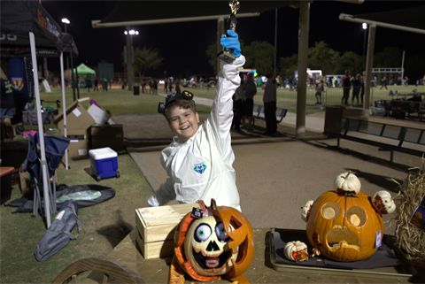 A child in a white costume joyfully holds up a trophy next to creatively carved pumpkins with spooky faces at an outdoor nighttime event.