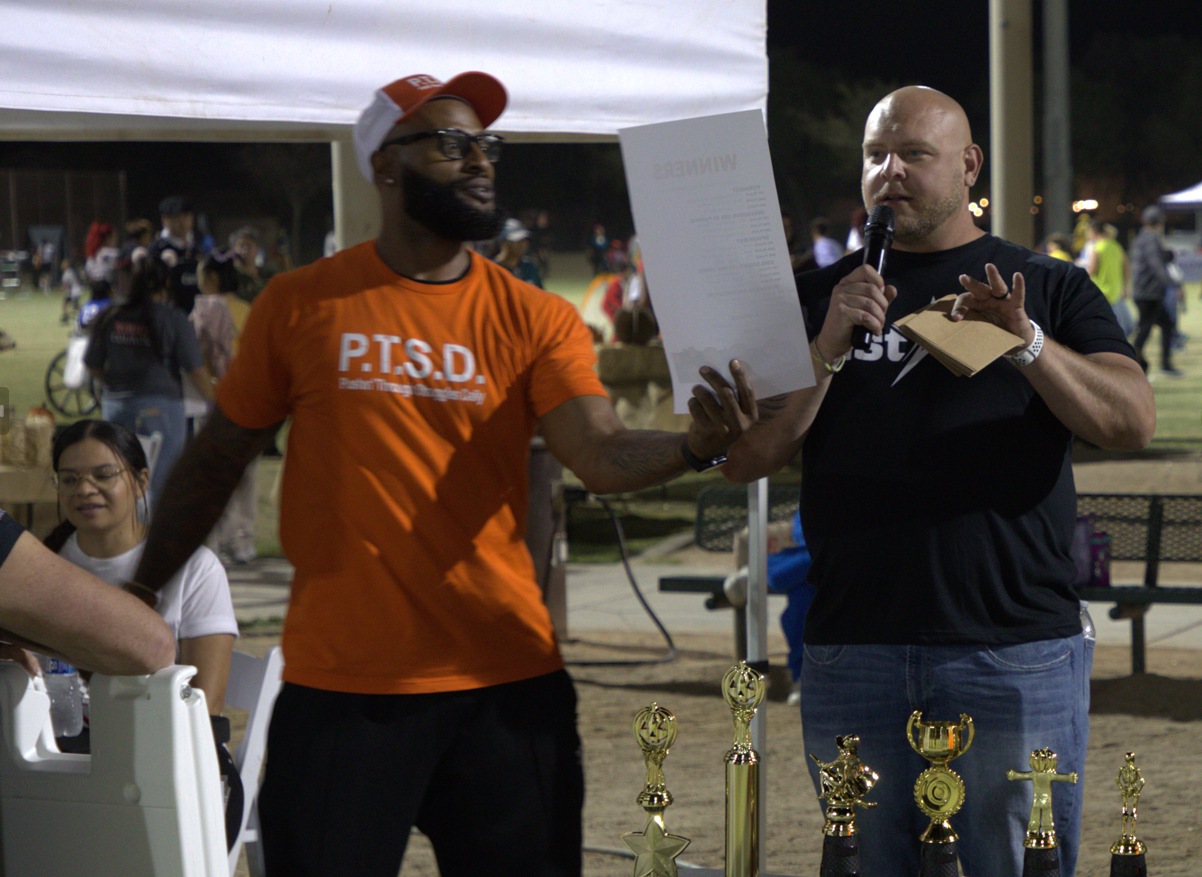 Two men stand under a white tent at an outdoor event. One wears an orange shirt with 