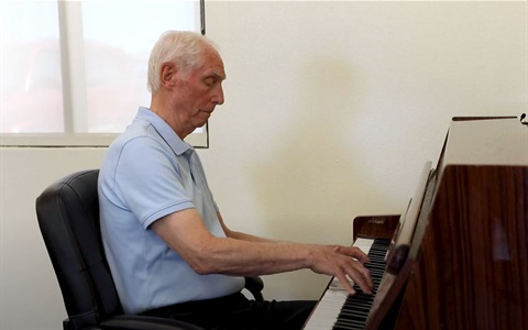 A man with short white hair plays the piano intently in a simple, light-colored room, creating a calm and focused atmosphere.