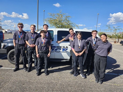A group of eight young individuals wearing matching dark uniforms stand proudly in front of a police vehicle. Many have medals around their necks, conveying achievement and teamwork. The sky is clear and blue, suggesting a sunny day.