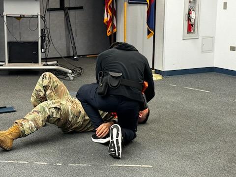 A person in a military uniform and a man in a black jacket lie on a gray carpeted floor, simulating a tactical drill. Flags and equipment are in the background.
