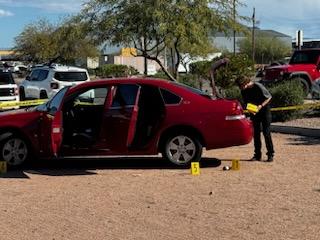 A red car is parked on gravel with doors open, surrounded by police tape and evidence markers. A person examines the scene under a clear blue sky.