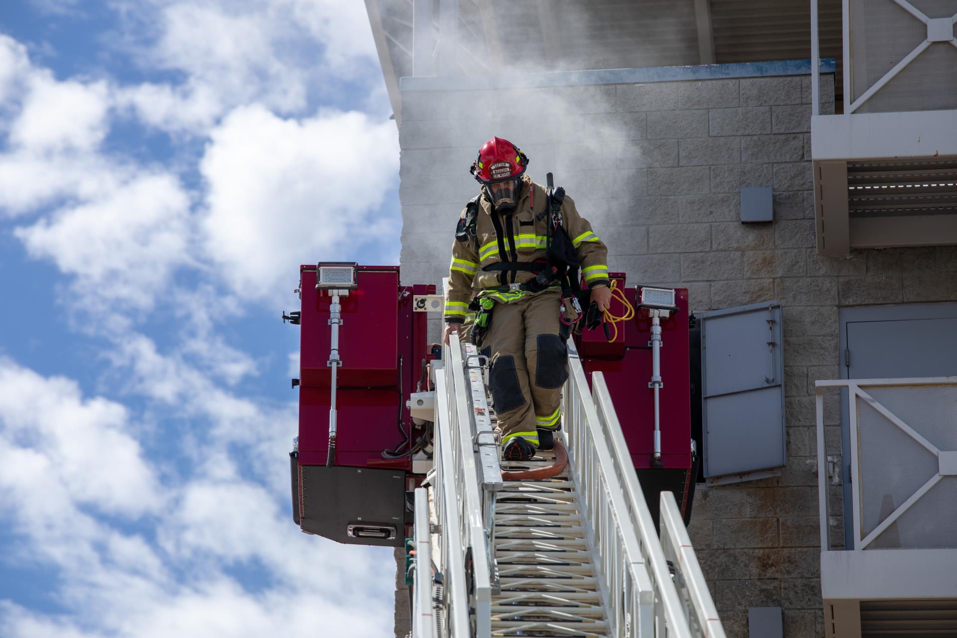 Firefighter in full gear descends a ladder from a smoky building. Bright blue sky with clouds in the background, scene conveys urgency and bravery.