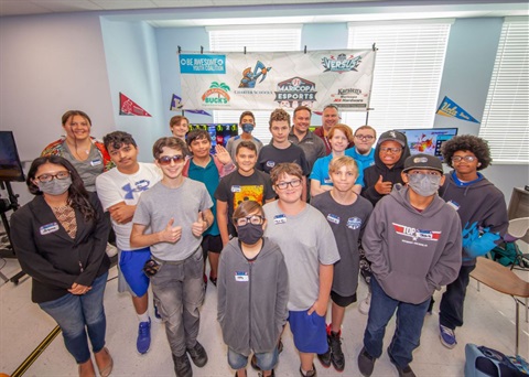 A diverse group of smiling teens and adults pose together in a well-lit room, surrounded by banners related to esports and schools. The mood is upbeat and friendly.