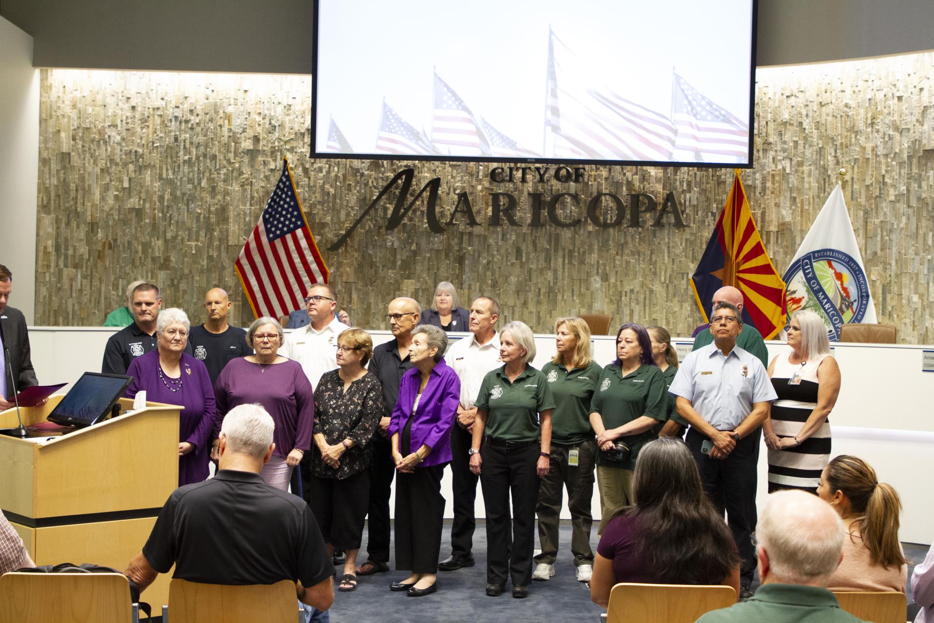 A group of people stand together inside the City of Maricopa council chamber. American, Arizona state, and city flags are displayed in the background.