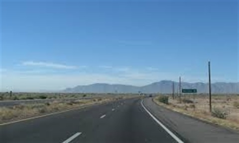 A long, empty highway stretches under a clear blue sky, flanked by flat, arid desert and distant mountains, conveying a sense of solitude and vastness.
