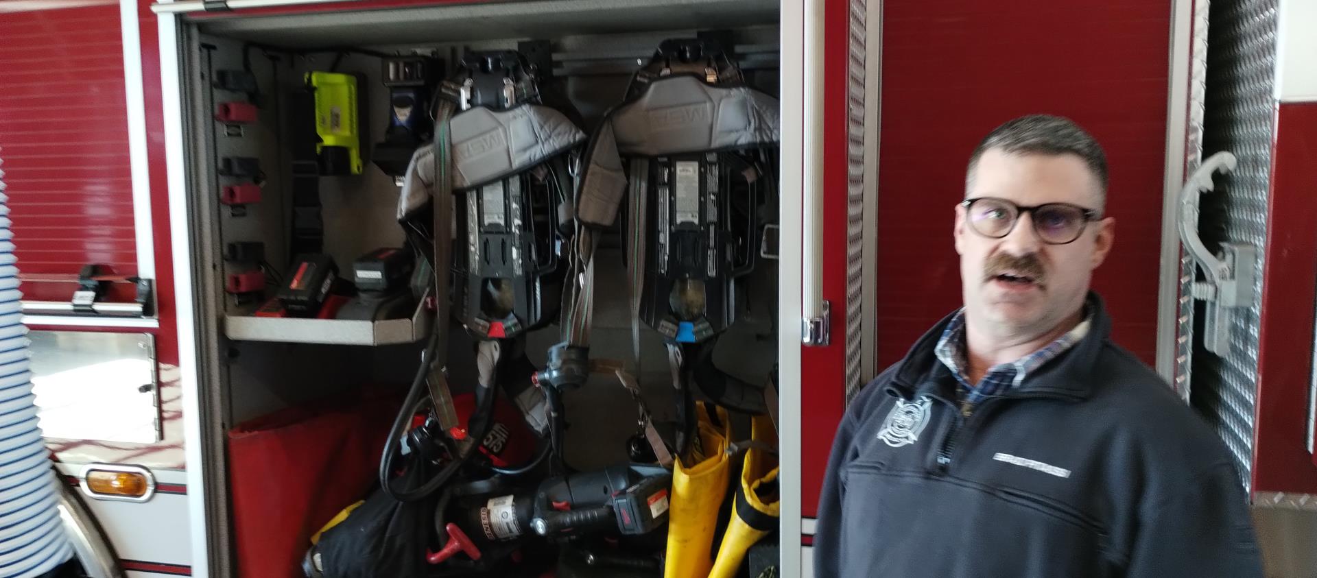 A firefighter with a mustache stands beside an open fire truck compartment filled with equipment, including two breathing apparatuses and various tools.