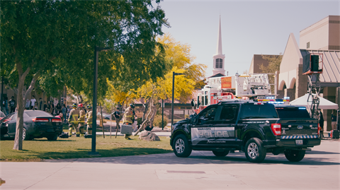 Police and fire vehicles are at a suburban scene with emergency personnel near a damaged car under trees. A church steeple is visible in the background.