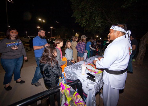 A man in a karate costume distributes candy to a line of children at a nighttime event. The scene is lively as families gather around, enjoying the festivities.