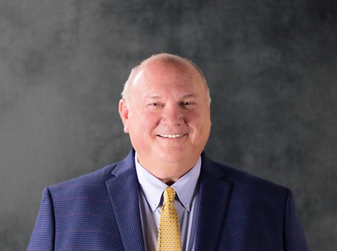 A smiling man in a blue suit and yellow tie stands against a dark, textured background. He conveys a professional and welcoming demeanor.