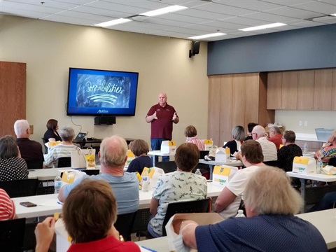 A man in a maroon polo gives a presentation to a classroom of seated adults. A screen displays 