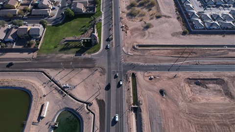 Aerial view of a suburban T-junction with sparse traffic. Surrounding areas include residential homes with green lawns, dry dirt lots, and a canal. Sunny day.