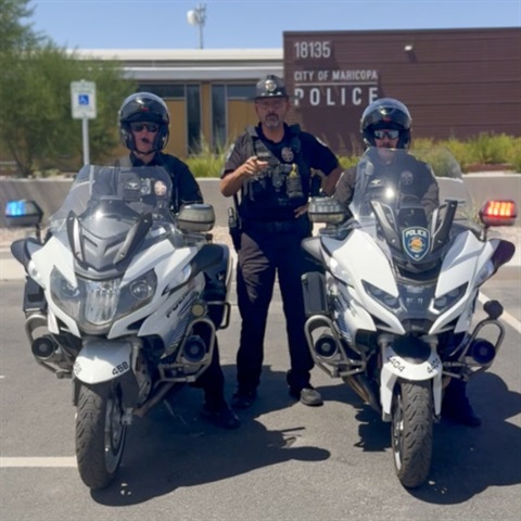 Two police officers sit on white motorcycles with their helmets on, flanking another officer standing between them. They are in front of a police station.