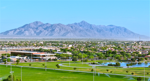 A sprawling cityscape with suburban houses is set against distant, rugged mountains under a clear blue sky. In the foreground, lush green parks and a reflective pond provide a tranquil, scenic contrast.