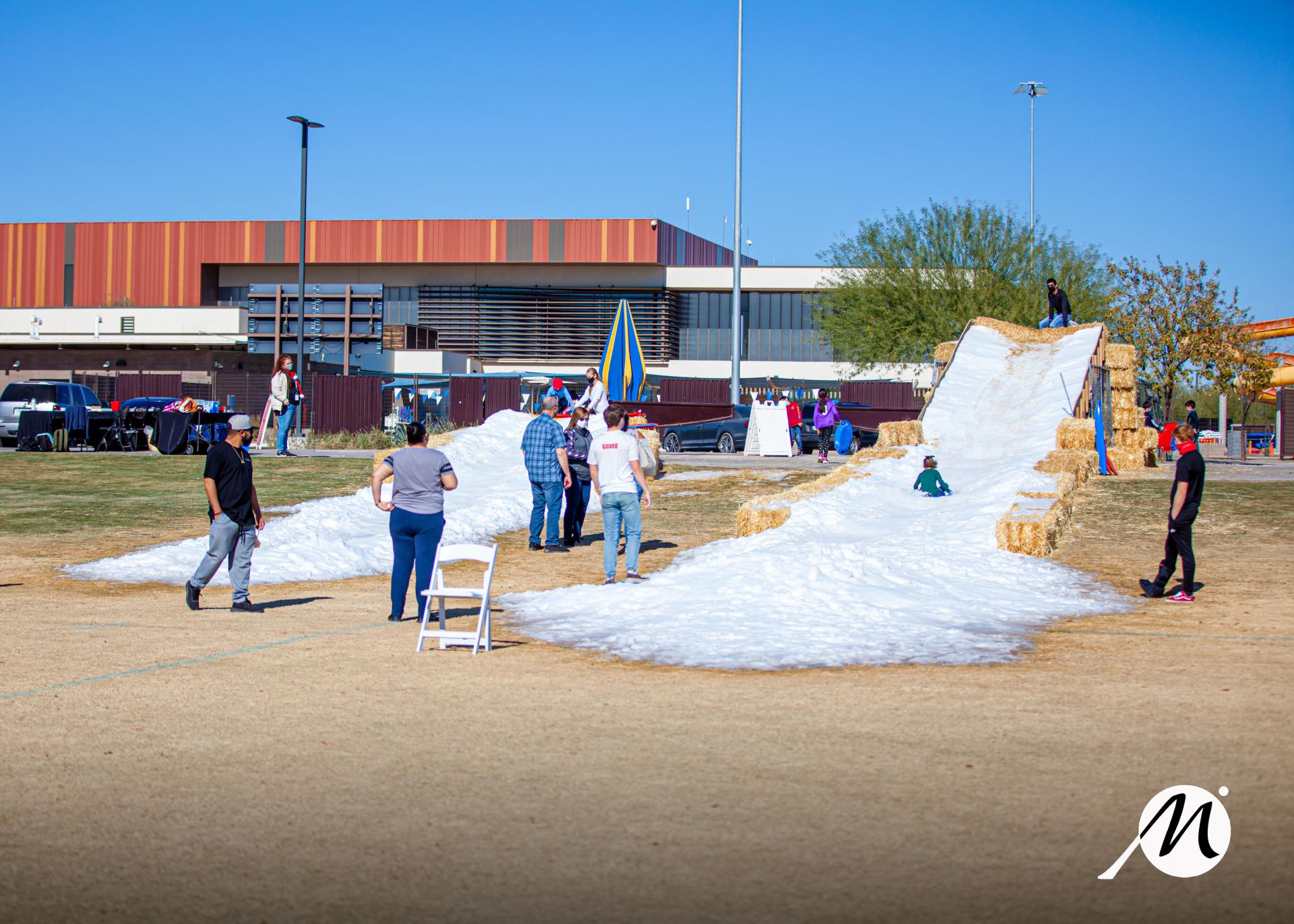 Outdoor scene with people enjoying a snow slide made of hay bales covered in snow. A large building and a tree are in the background, under a clear blue sky.