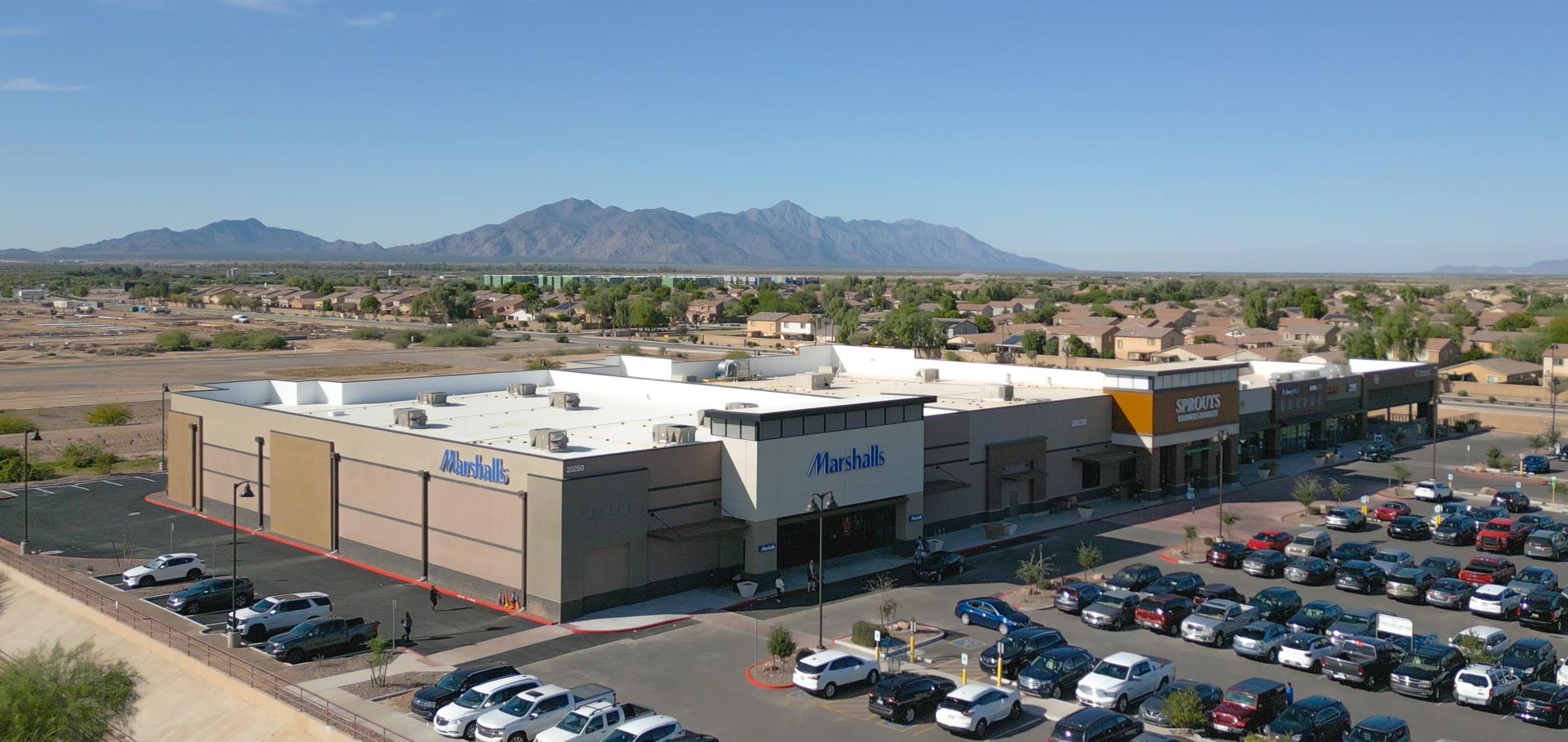 Aerial view of a shopping center with several retail stores, including Marshalls. The parking lot is full, and mountains are visible in the background.