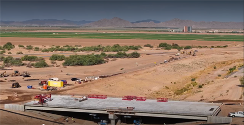 A vast desert landscape with a partially constructed bridge in the foreground. Construction vehicles are scattered along the sandy terrain. In the distance, fields, a large building, and mountains are visible under a clear blue sky.